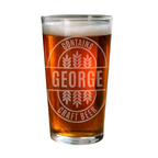 A clear pint glass filled with amber craft beer and topped with white foam, featuring a frosted circular logo that reads 'CONTAINS GEORGE CRAFT BEER' with wheat illustrations, on a white background.