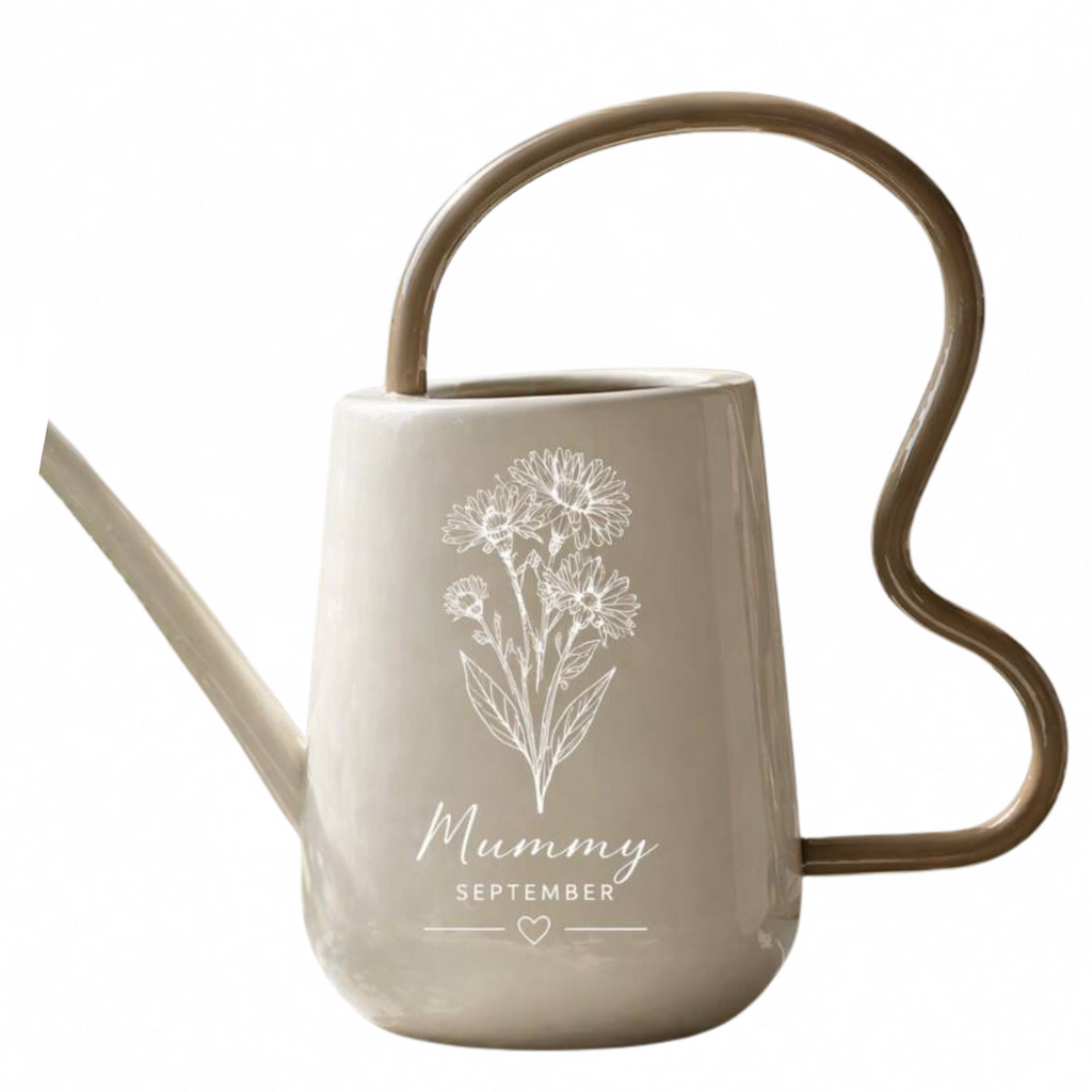 A ceramic watering can with a matte beige body, featuring a white line-art floral design and the text 'Mummy SEPTEMBER' on the front. It has a curved metallic gold handle and a long spout, set against a plain white background.