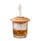 A whiskey glass with a wooden lid engraved with 'DAD', filled with amber liquor and ice, with white smoke rising from the top against a white background.