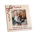 A square cream-colored ceramic photo frame with a red ribbon design in the top-left corner and the text 'THE WALKER FAMILY OUR HOME FILLED WITH LOVE' at the bottom. The frame is shown displaying a family photo on a clean white background.