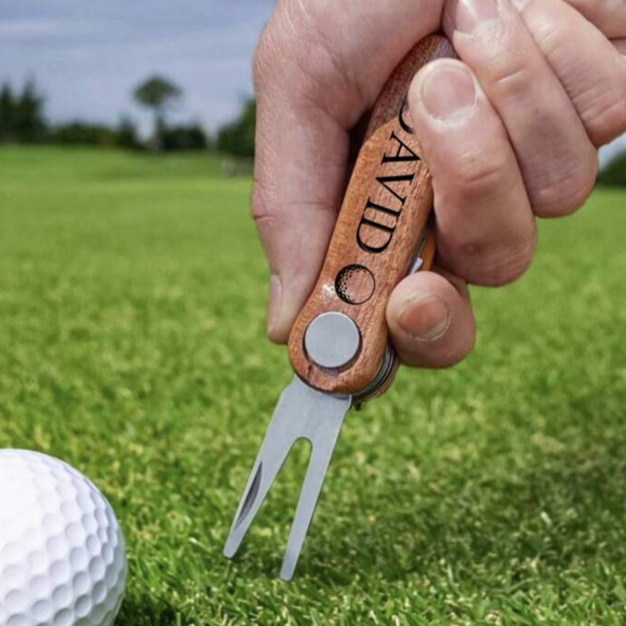 Hand holding a golf divot tool next to a golf ball on a green field