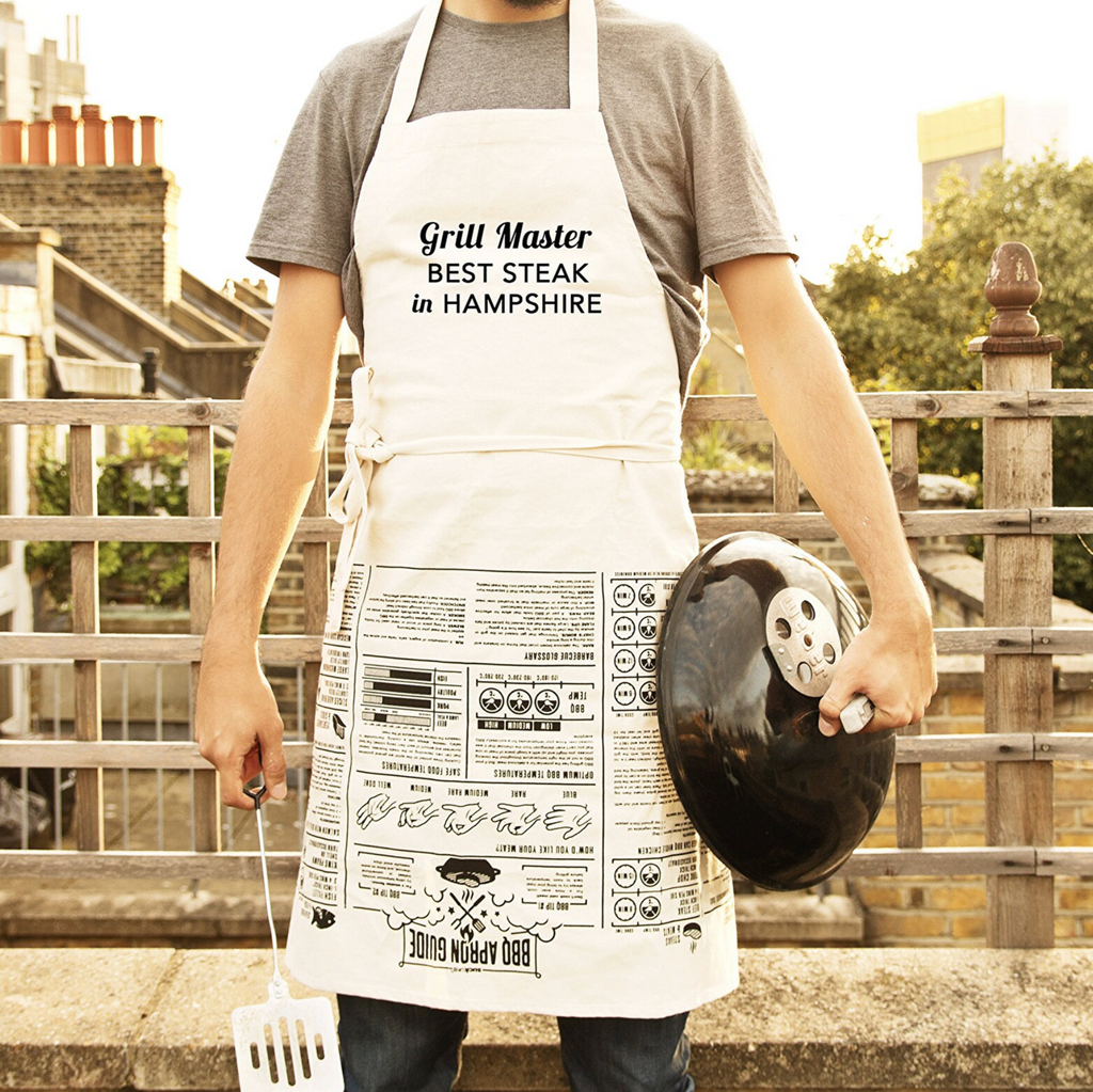 Person wearing a 'Grill Master BEST STEAK in HAMPSHIRE' apron holding a barbecue lid outdoors.
