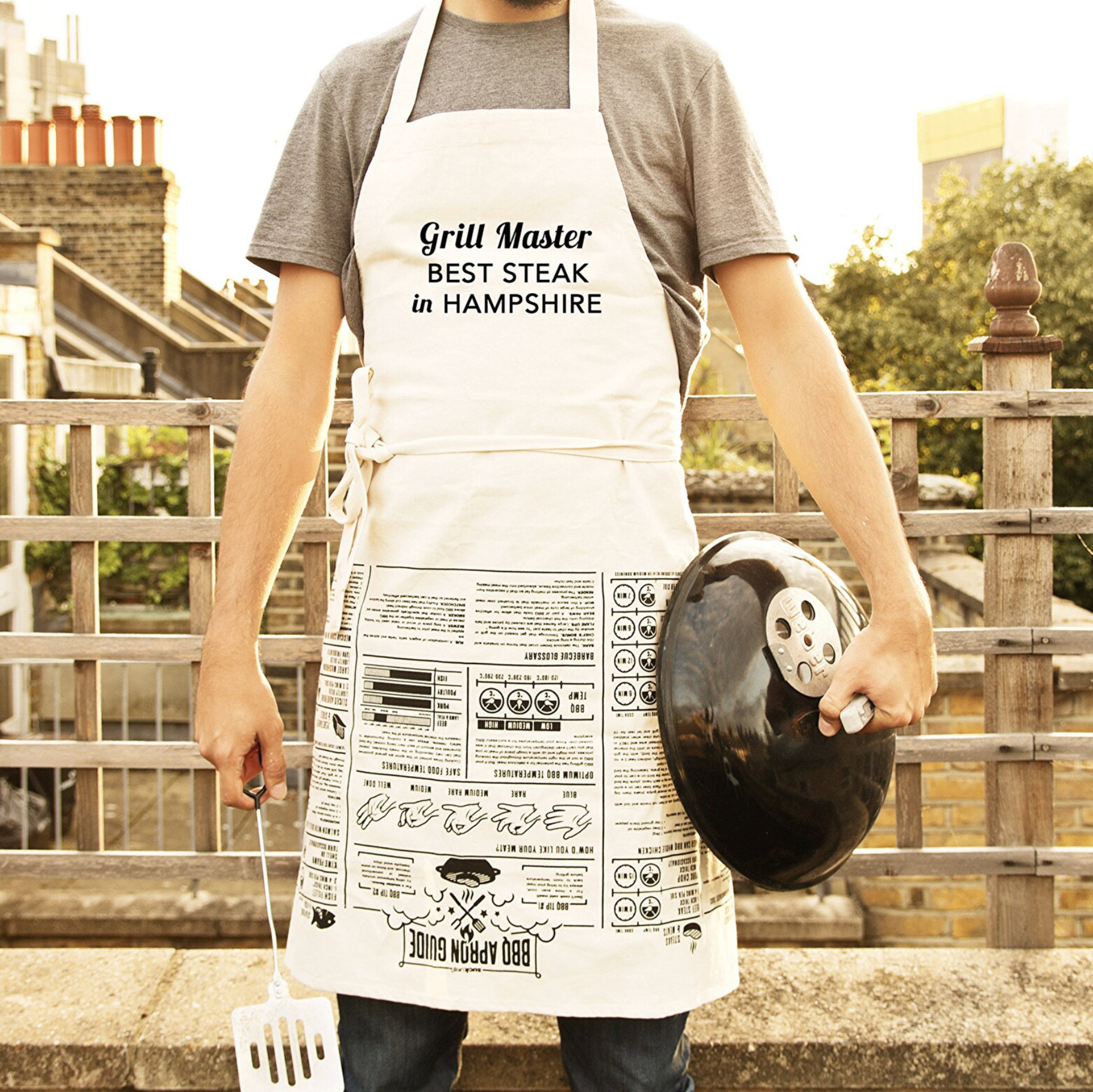 Person wearing a 'Grill Master BEST STEAK in HAMPSHIRE' apron holding a barbecue lid outdoors.