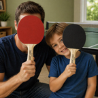 Man and child holding ping pong paddles in front of their faces in a room with a ping pong table.