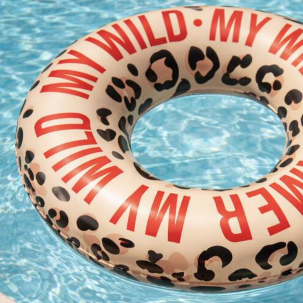 An inflatable swim ring floating in a swimming pool, featuring a beige leopard print pattern with bold red text reading 'MY WILD SUMMER' and black leopard spots. A person's feet with red toenail polish and rings are visible at the bottom edge of the pool.