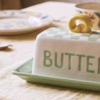 A ceramic butter dish with a white lid featuring a light green checkerboard pattern and the word 'BUTTER' in green, topped with a small yellow handle. The dish sits on a matching green tray on a beige tablecloth, with a floral jar in the background.