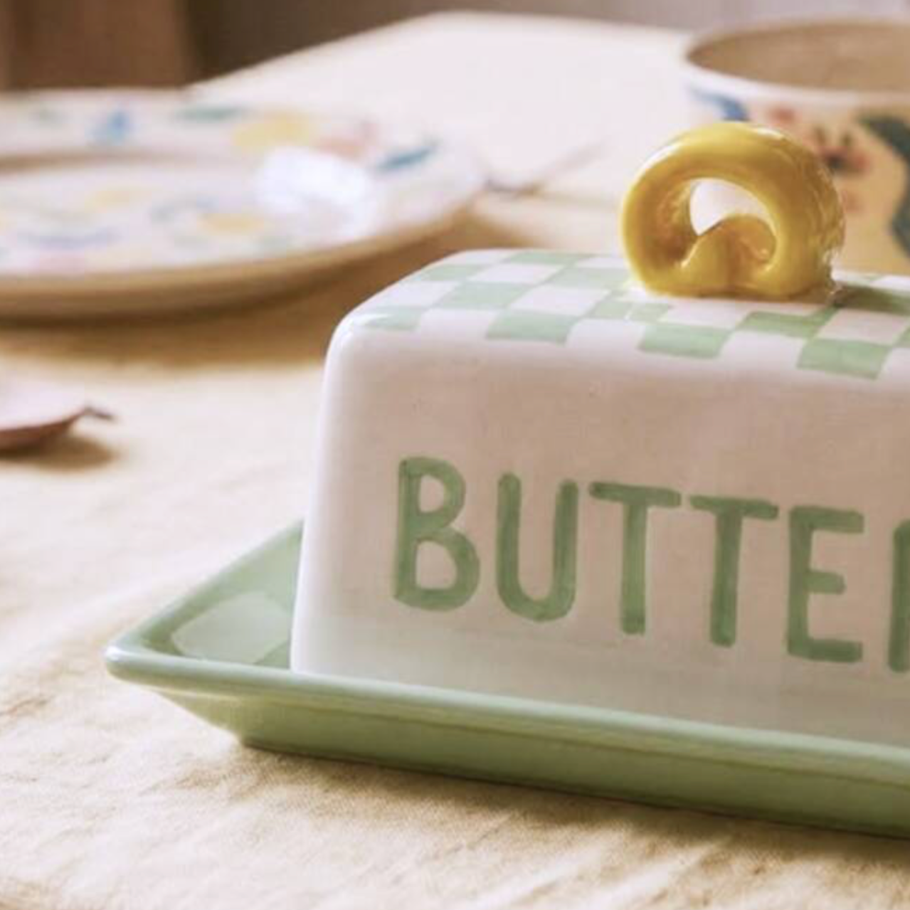A ceramic butter dish with a white lid featuring a light green checkerboard pattern and the word 'BUTTER' in green, topped with a small yellow handle. The dish sits on a matching green tray on a beige tablecloth, with a floral jar in the background.
