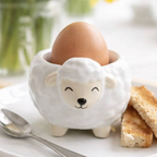 A white ceramic lamb-shaped egg holder with a brown egg inside, styled on a white plate with toast and a spoon, with yellow daffodils in the background.