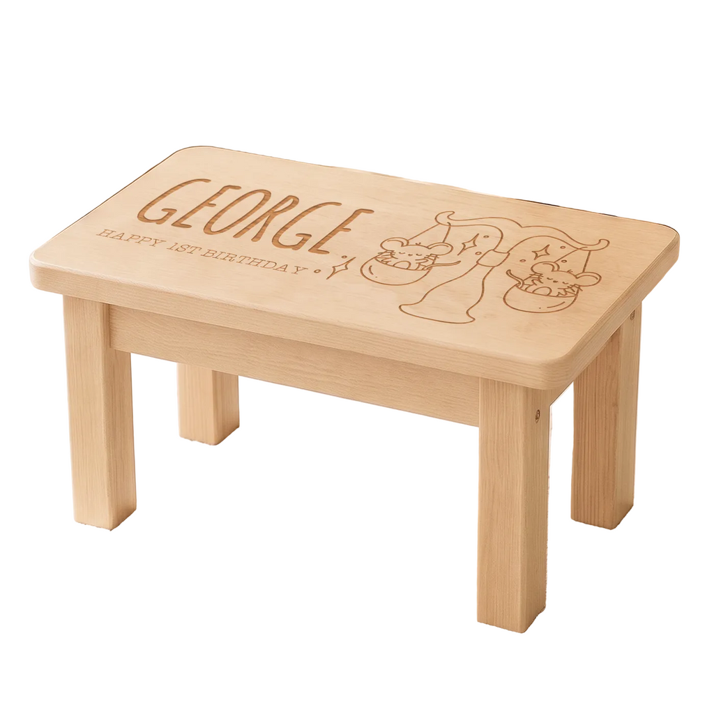 A small light wood children's stool or bench with engraved personalization reading 'GEORGE' and 'HAPPY 1st BIRTHDAY' plus cartoon characters, shown in a styled nursery setting on a star-patterned rug.