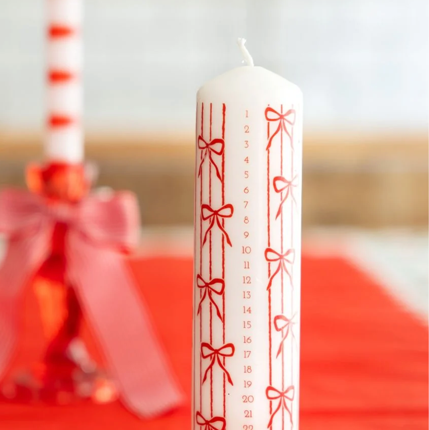 A white taper candle with red ribbon and bow illustrations and numbered vertical lines, standing on a red fabric surface. A second blurred candle with red stripes and a pink bow sits in the background.