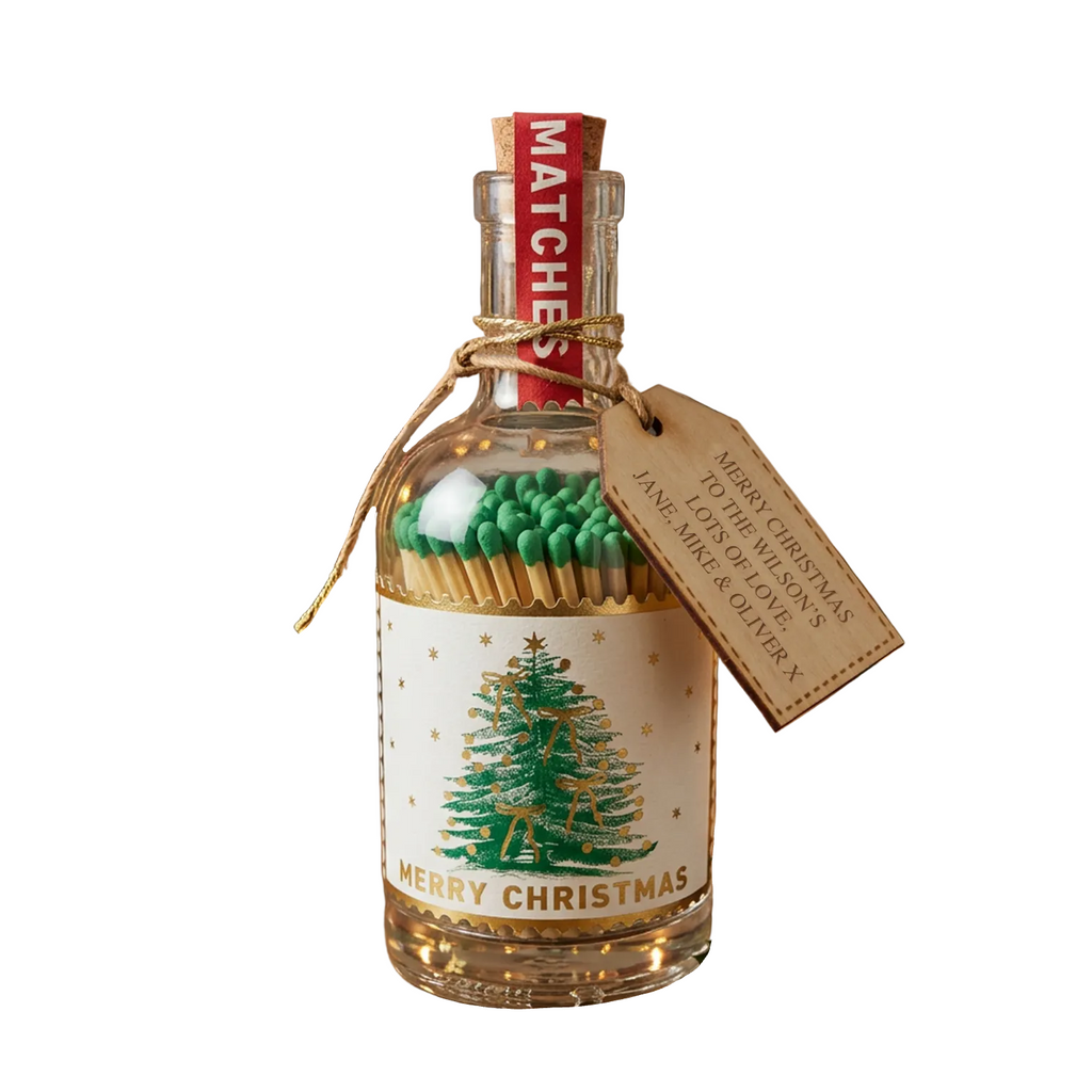 A Christmas-themed glass bottle filled with green matches and topped with a red matchstick cap, wrapped in twine with a wooden tag that reads 'Merry Christmas to the gift of your life, Jane aka Olivera.' The bottle is set on a wooden table with a blurred festive background featuring candles, pine branches, and a lit fireplace.