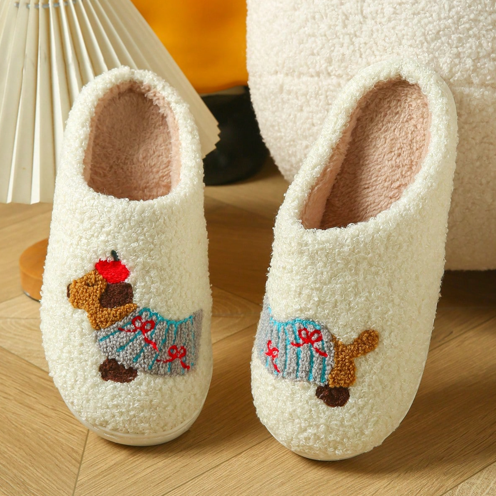 A pair of cream sherpa slip-on house slippers with embroidered dachshund dogs wearing blue sweaters, displayed on a wooden floor next to a lamp.