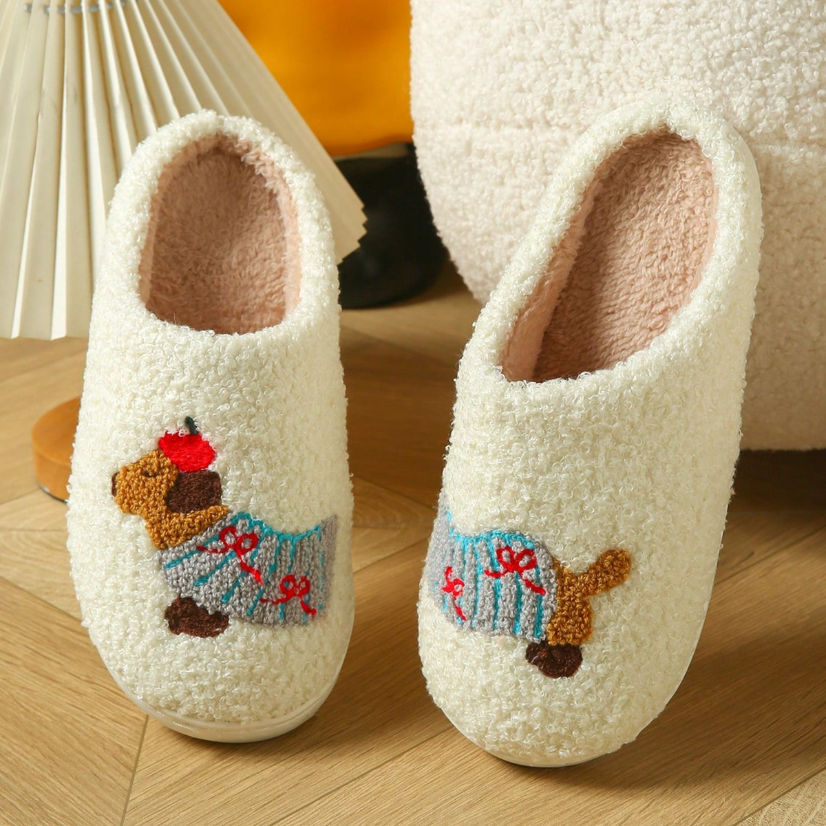A pair of cream sherpa slip-on house slippers with embroidered dachshund dogs wearing blue sweaters, displayed on a wooden floor next to a lamp.