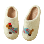 A pair of cream sherpa slip-on house slippers with embroidered dachshund dogs wearing blue sweaters, displayed on a wooden floor next to a lamp.