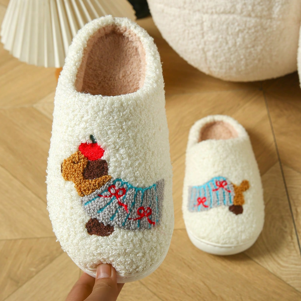 A pair of fluffy cream-colored indoor slippers with embroidered dachshund designs wearing blue sweaters and red bows, shown on a wooden floor with a hand holding one slipper.