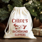 A cream-colored fabric drawstring gift pouch printed with 'CHLOE'S DACHSHUND SLIPPERS' and a dachshund illustration in dark red, set against a festive Christmas tree background.