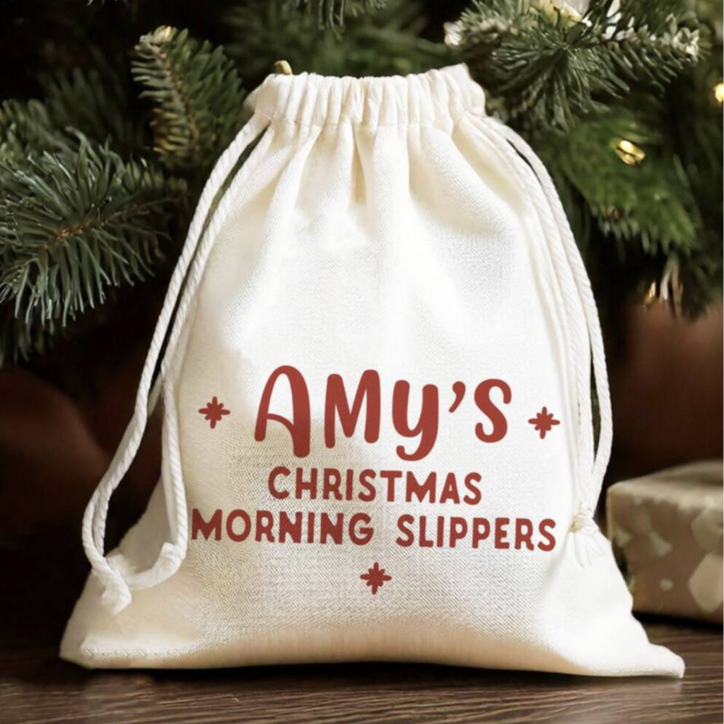 An off-white cotton Christmas stocking bag with red printed text reading 'Amy's Christmas Morning Slippers' sits on a wooden surface in front of a Christmas tree.
