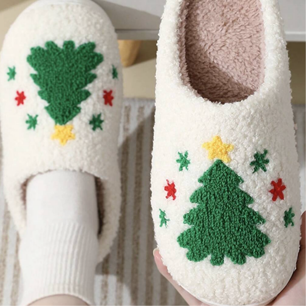 A pair of fluffy white slip-on house slippers with a green Christmas tree and small red and yellow star pattern, shown on a foot and held in a hand against a soft neutral background.