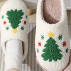 A pair of fluffy white slip-on house slippers with a green Christmas tree and small red and yellow star pattern, shown on a foot and held in a hand against a soft neutral background.