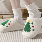 A person wearing white fuzzy slip-on slippers with embroidered Christmas trees and small holiday motifs, paired with white socks in a cozy indoor setting.