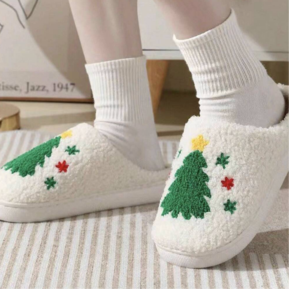 A person wearing white fuzzy slip-on slippers with embroidered Christmas trees and small holiday motifs, paired with white socks in a cozy indoor setting.