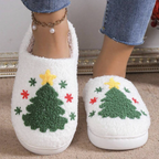 A person wearing white fuzzy slip-on slippers with a green Christmas tree design, red and yellow accents, and blue jeans, standing on a beige carpet.