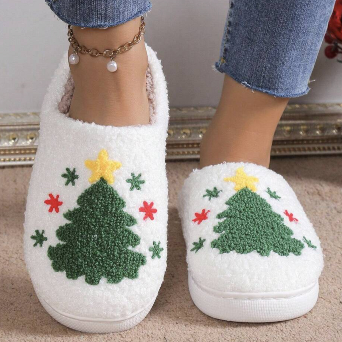 A person wearing white fuzzy slip-on slippers with a green Christmas tree design, red and yellow accents, and blue jeans, standing on a beige carpet.