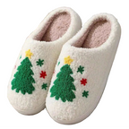 A pair of white sherpa slip-on house slippers with a green Christmas tree and colorful embroidered stars on the side, displayed on a striped fabric surface.