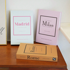 Three minimalist hardcover books titled 'Little Book of [City]' in white, pale pink, and tan are arranged on a wooden table against a light wall.
