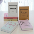 Five hardcover books titled 'Little Book of [City Name]' arranged on a white surface in front of a softly lit window. The books include Berlin, Rome, Amsterdam, Tokyo, and Milan, with different cover colors: white, tan, pink, beige, and light blue.