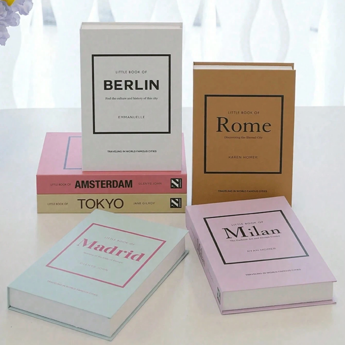 Five hardcover books titled 'Little Book of [City Name]' arranged on a white surface in front of a softly lit window. The books include Berlin, Rome, Amsterdam, Tokyo, and Milan, with different cover colors: white, tan, pink, beige, and light blue.