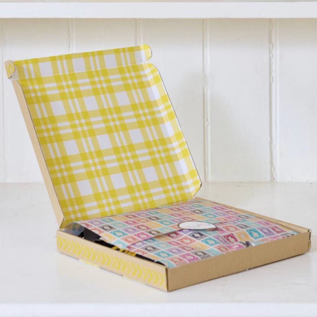 An open cardboard gift box with a yellow and white plaid patterned lining and a colorful patterned insert, set on a white shelf against a white paneled wall.