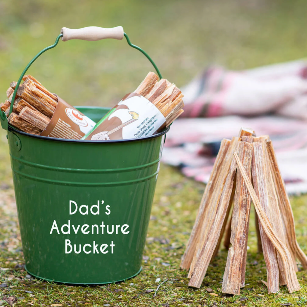 Green bucket labeled 'Dad's Adventure Bucket' with firewood and a small wooden campfire structure outdoors.