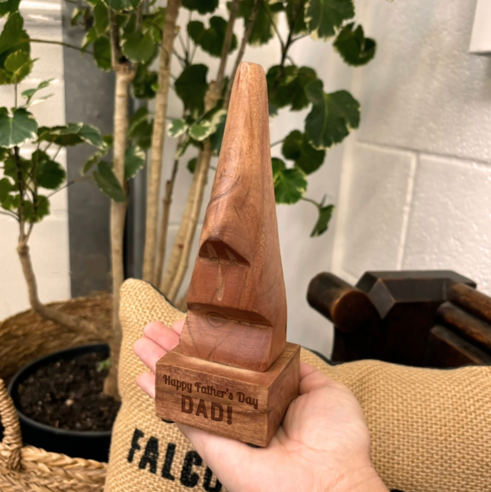 Wooden trophy with 'Happy Father's Day DAD!' engraving held by a person, with plants and a wall in the background.