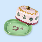 Hand lifting the domed lid of the Pink And Green Cactus Butter Dish to reveal a block of butter on the green dish