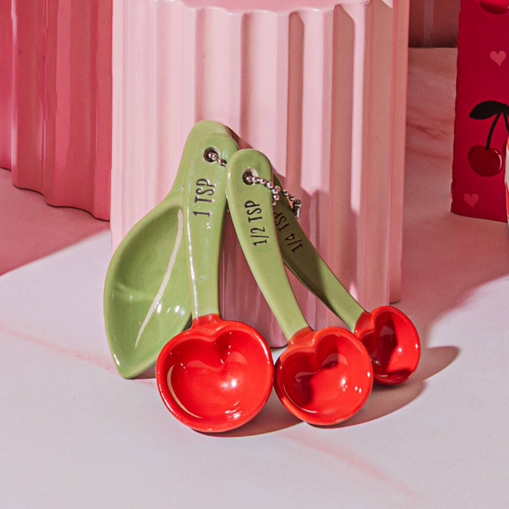 Cherry Ceramic Measuring Spoons leaning against a pink ribbed column showing the red cherry bowls and green handles