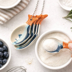 Sardine Ceramic Measuring Spoons hanging on silver ball chain on a marble surface next to blueberries and rosemary with one spoon scooping sugar
