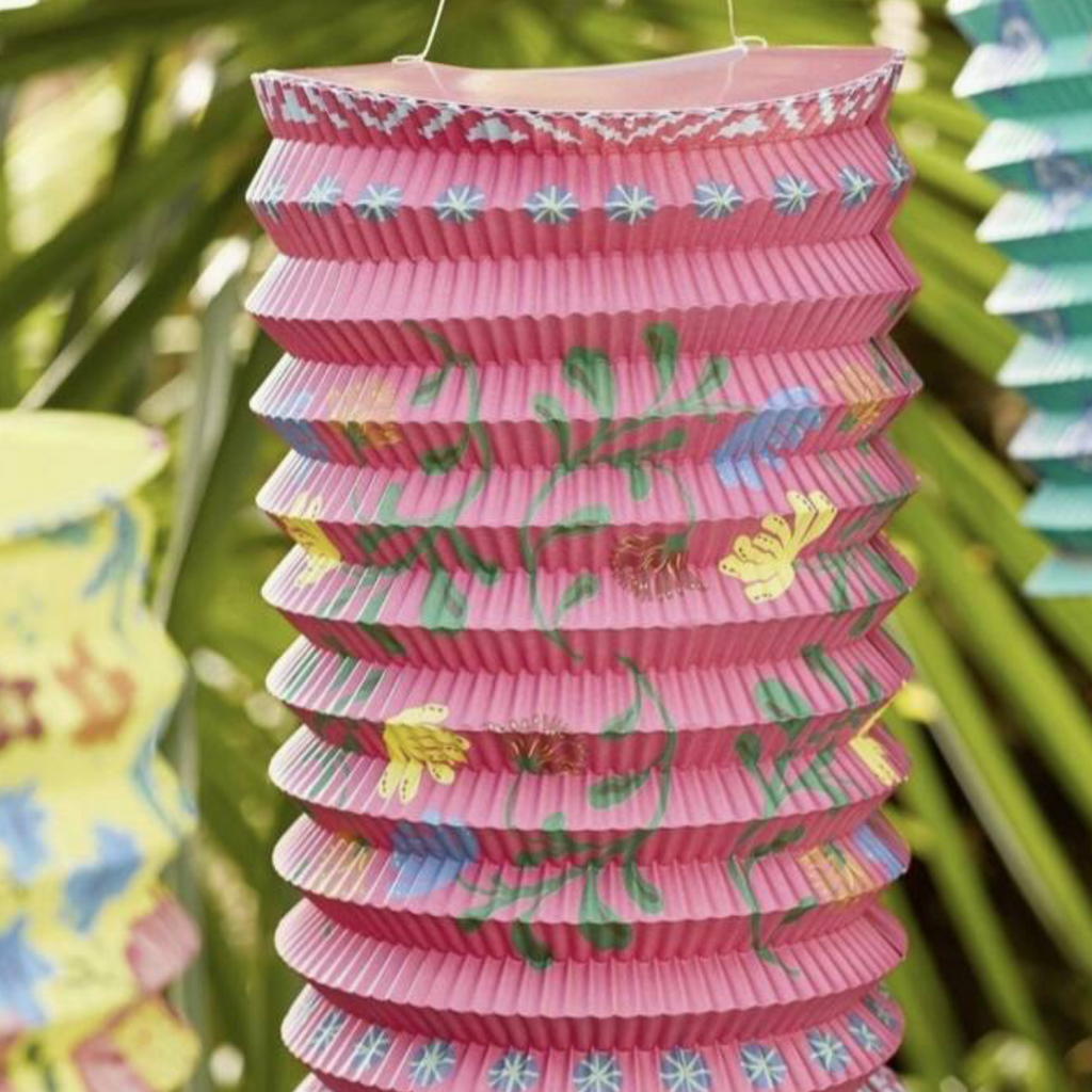 Pink paper lantern with teal tassel hanging among yellow and teal lanterns in a tropical garden setting