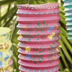 Pink paper lantern with teal tassel hanging among yellow and teal lanterns in a tropical garden setting