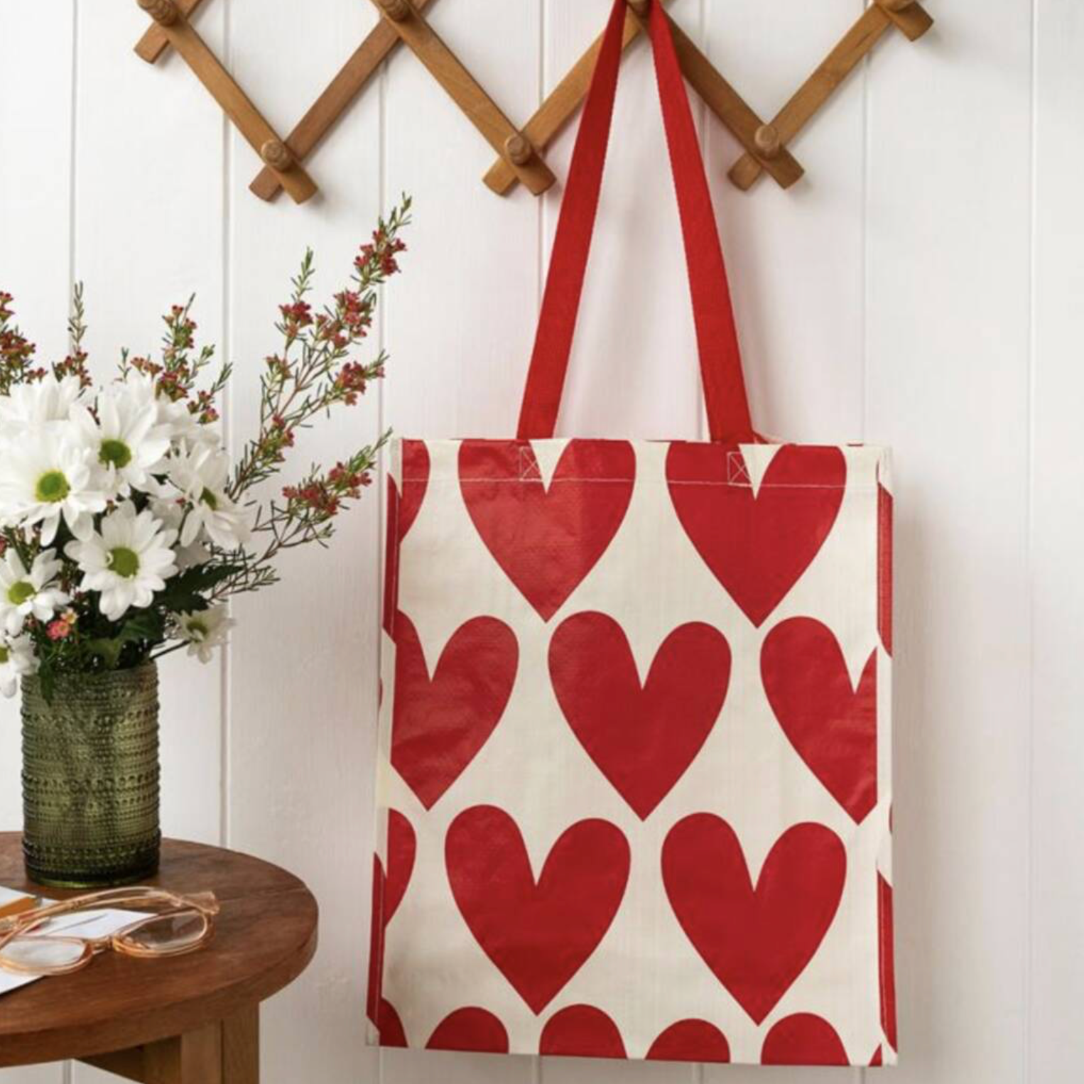 Close up of Recycled Hearts Shopping Bag leaning against a white panelled wall with white daisies and red flowers in background