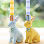 Hand Painted Bunnies Ceramic Candle Holder pair styled on a wooden surface with yellow stripe and blue gingham dinner candles lit against a garden window