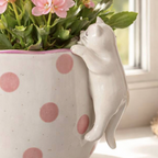 Close up of the white porcelain peeking cat on the Spotty Cat Planter showing the sleeping face and paws hooked over the rim with pink flowers