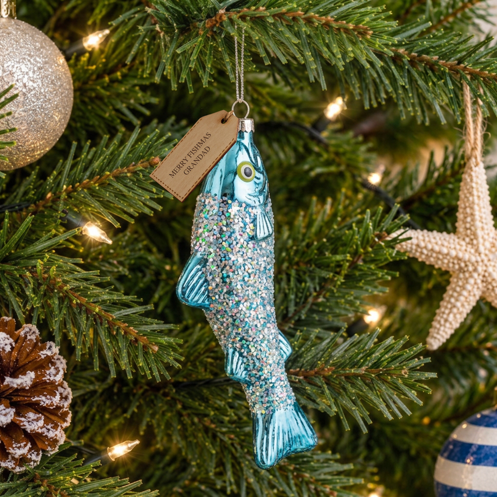 A fish-shaped Christmas ornament with blue glitter and a hanging tag reading 'MERRY CHRISTMAS GRANDPA,' displayed on a decorated Christmas tree with lights and other ornaments.