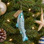 A fish-shaped Christmas ornament with blue glitter and a hanging tag reading 'MERRY CHRISTMAS GRANDPA,' displayed on a decorated Christmas tree with lights and other ornaments.
