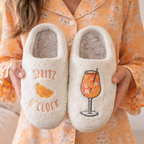 Cream Spritz O’Clock slippers with orange cocktail embroidery held up for display