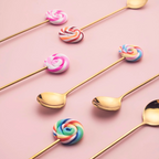 Gold lollipop spoons and forks with colourful handles arranged on a pink background