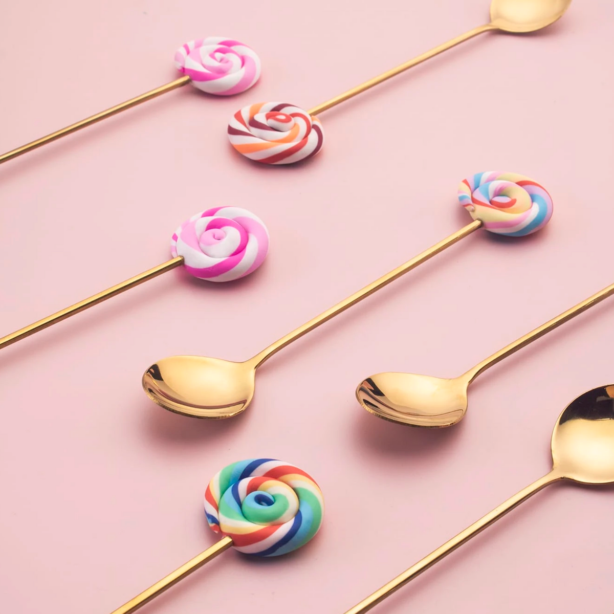 Gold lollipop spoons and forks with colourful handles arranged on a pink background