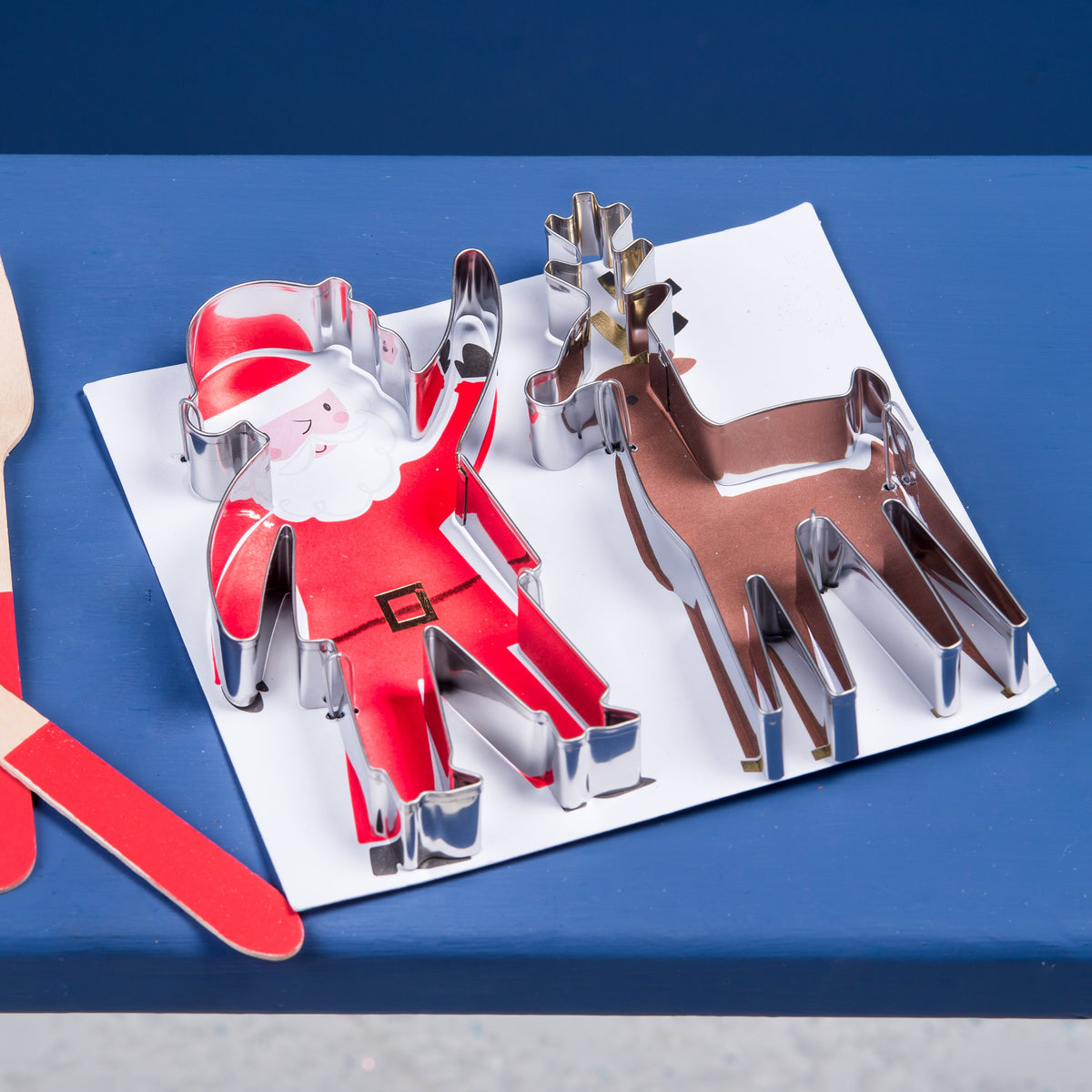 A set of Christmas-themed cookie cutters and wooden utensils arranged on a blue surface. The set includes a Santa Claus cutter, a reindeer cutter, a star cutter, and a snowman cutter on a white sheet, alongside a wooden spoon and fork with red handles.