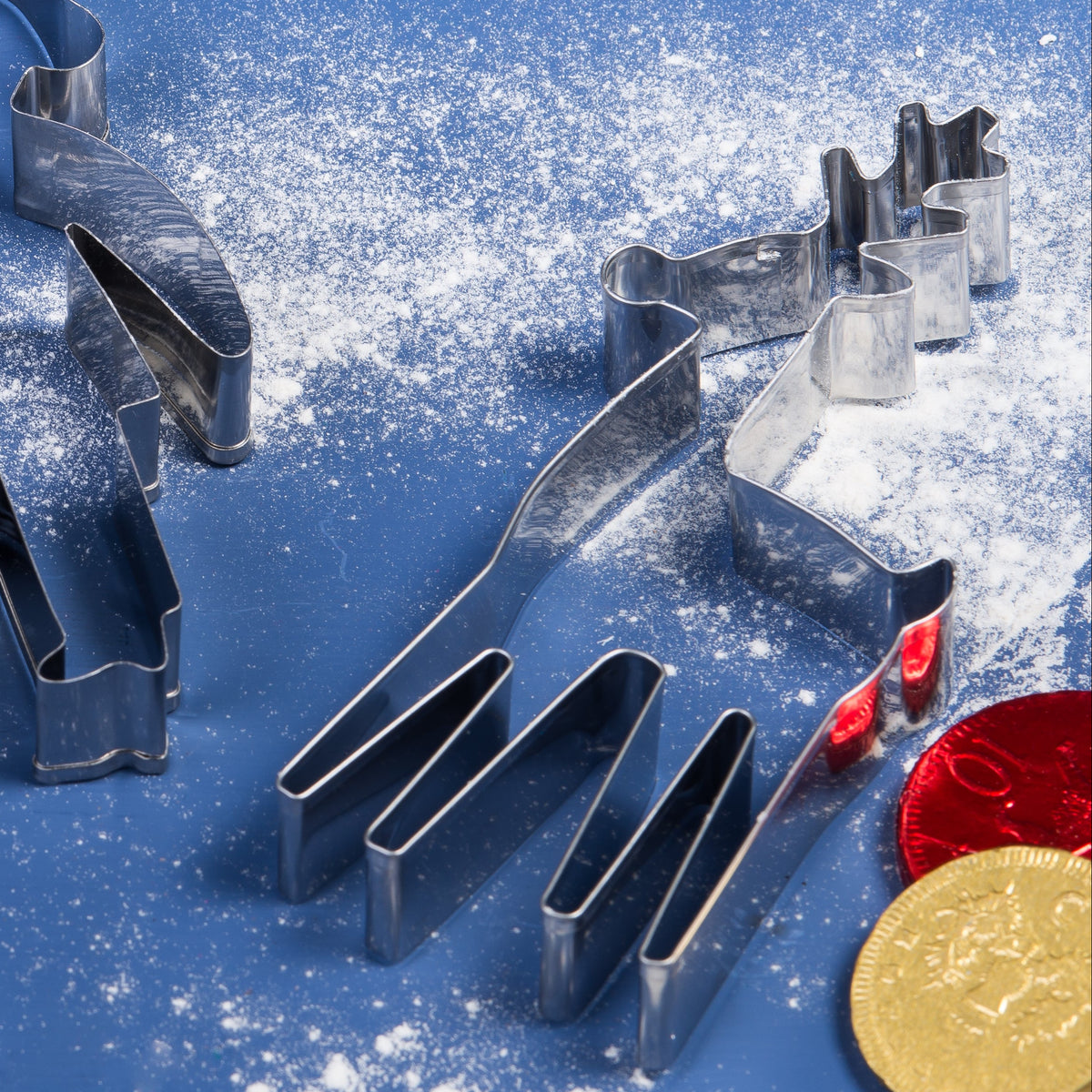 A set of metal holiday cookie cutters, including a reindeer and a penguin, arranged on a blue surface dusted with white flour, alongside wooden utensils with red handles and gold and red chocolate coins.