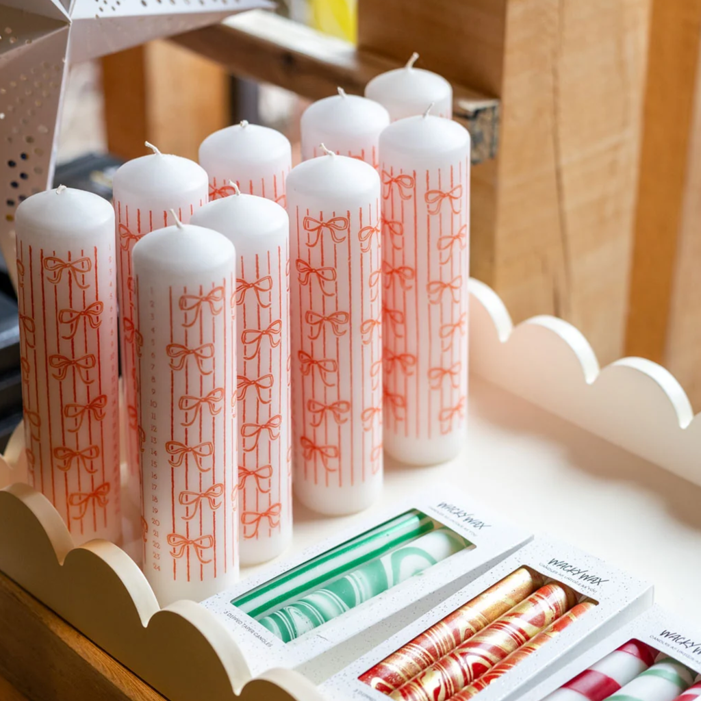 A set of white taper candles decorated with vertical red stripes and red bow motifs, displayed on a wooden shelf alongside packaged marbled and striped candles.